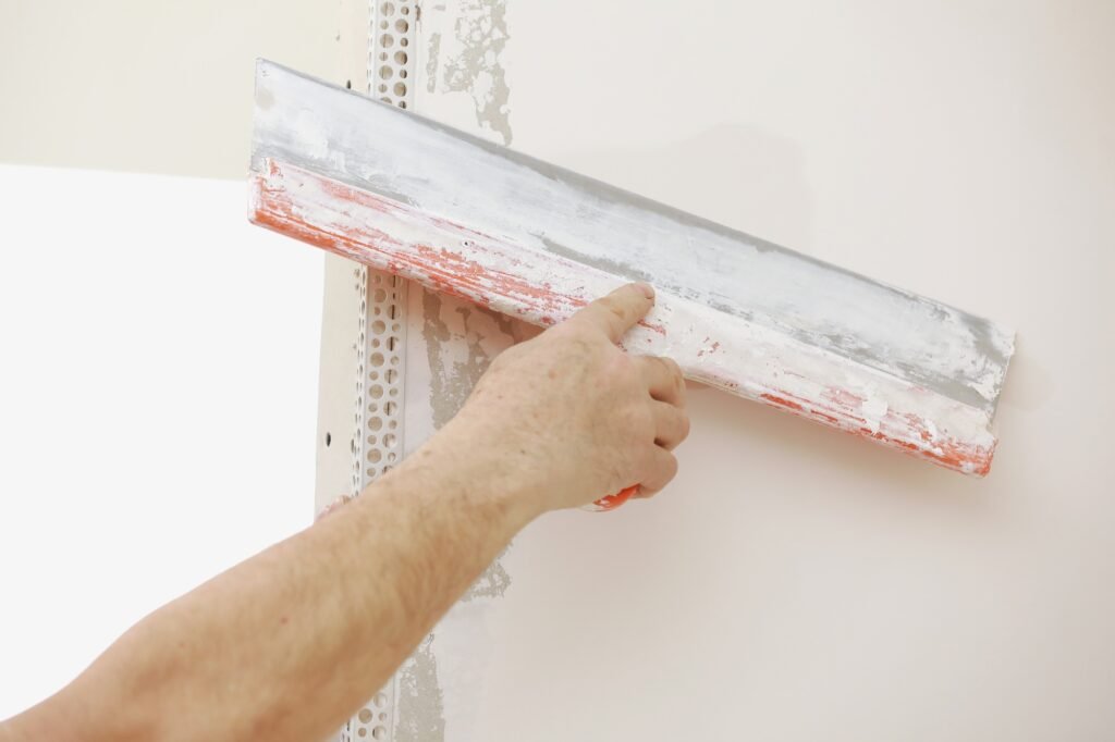 builder-in-protective-mask-using-a-trowel-to-add-plaster-plastering-wall-with-putty-knife-close